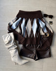 Brown argyle off-shoulder sweater styled with leather shorts, boots, and sunglasses