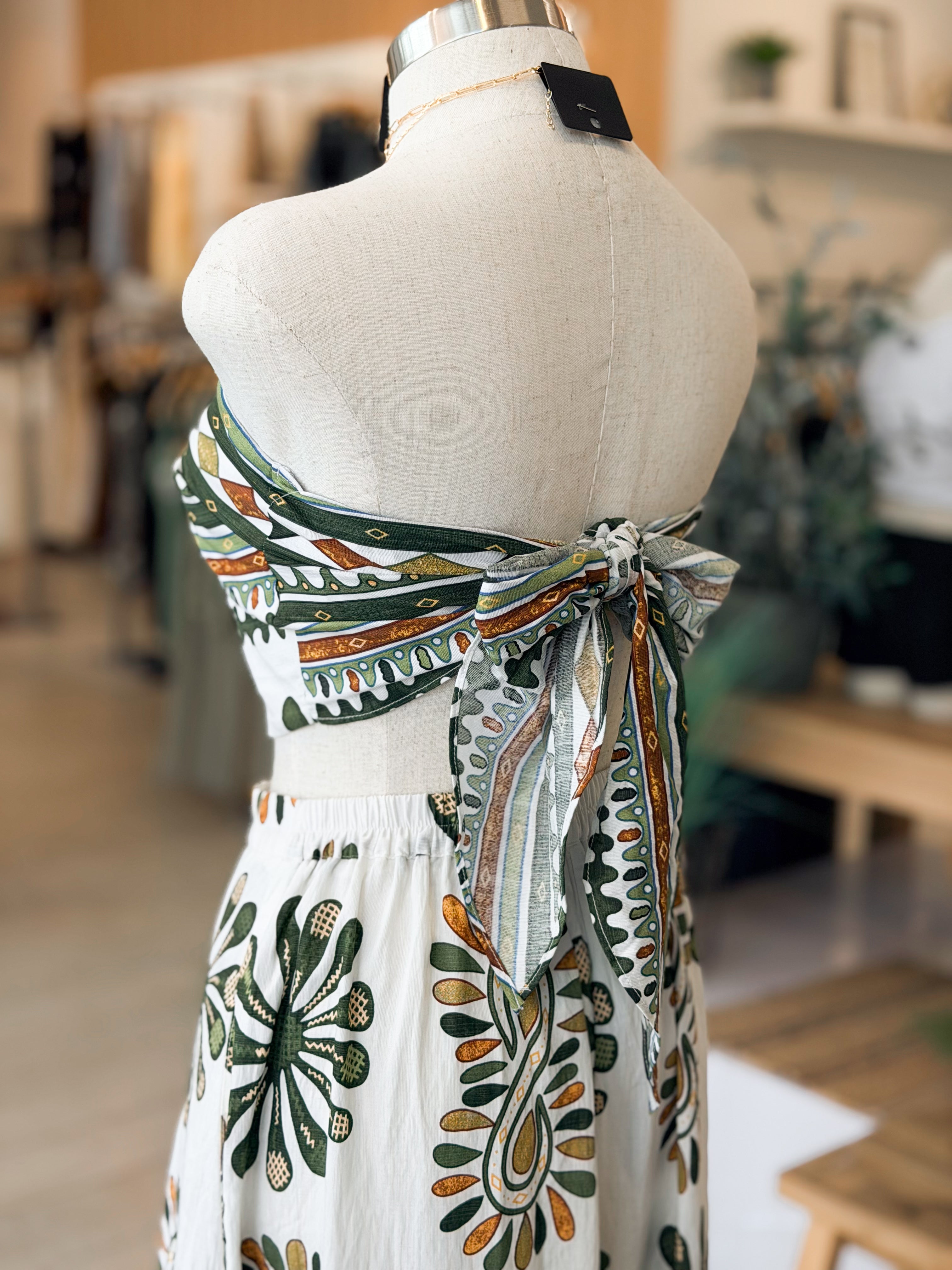 Mannequin wearing a white dress with green and orange floral patterns in a blurred indoor setting.