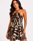 A woman modeling a halter-neck mini dress with a brown and beige zebra-like abstract print against a plain light gray studio background